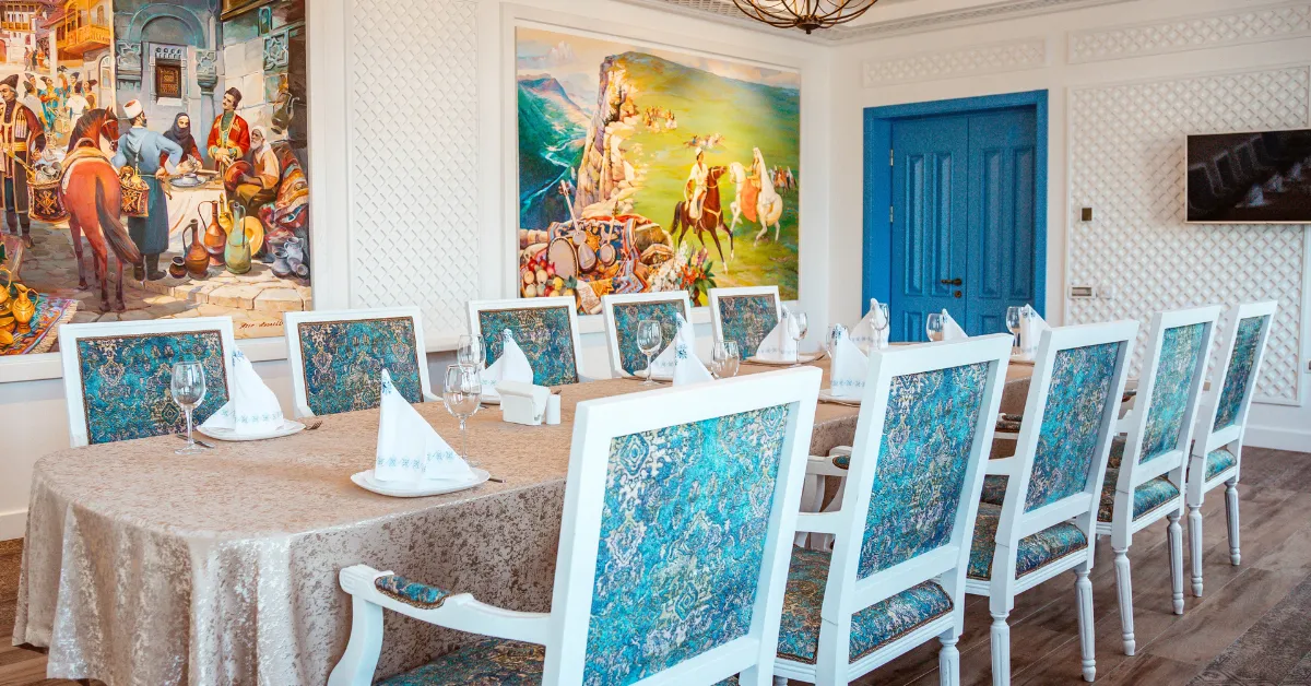 A stylish dining room with a long table set with folded napkins and glasses, surrounded by white chairs with blue patterned upholstery, colorful wall paintings, and a bright blue door.
