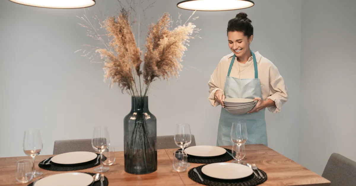 A woman wearing an apron placing plates on a wooden dining table set with glassware and black placemats, with a tall vase of dried pampas grass in the center under modern pendant lights.